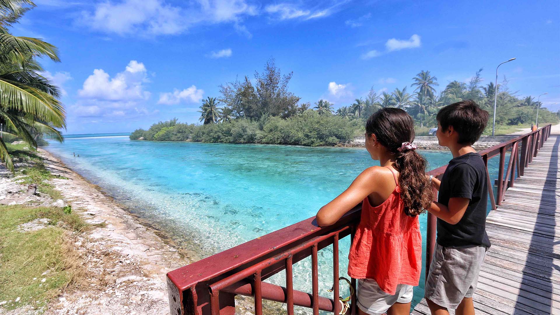 Two children standing on the wooden connecting bridge at Canareef Resort looking across the turquoise lagoon toward the neighboring local island of Hulhudhoo