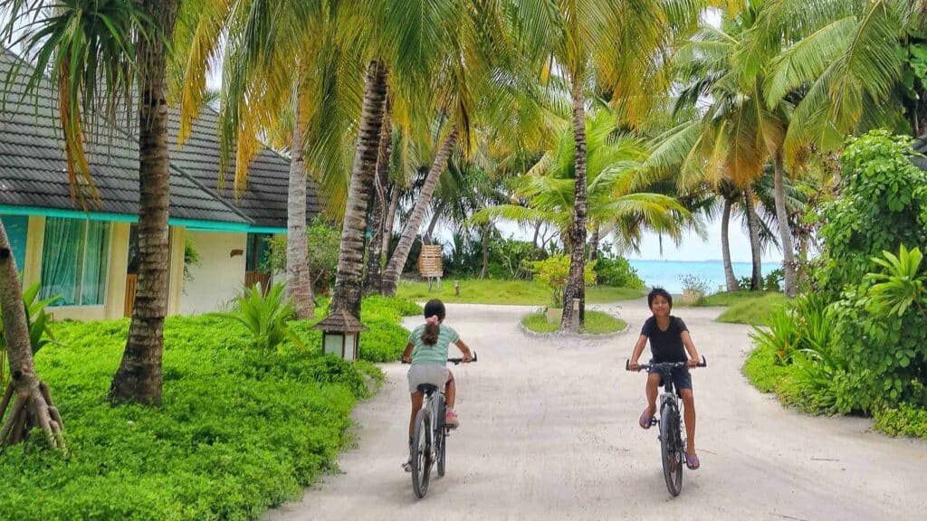 Two children riding bicycles along a lush, palm-fringed sand pathway near villas at Canareef Resort Maldives