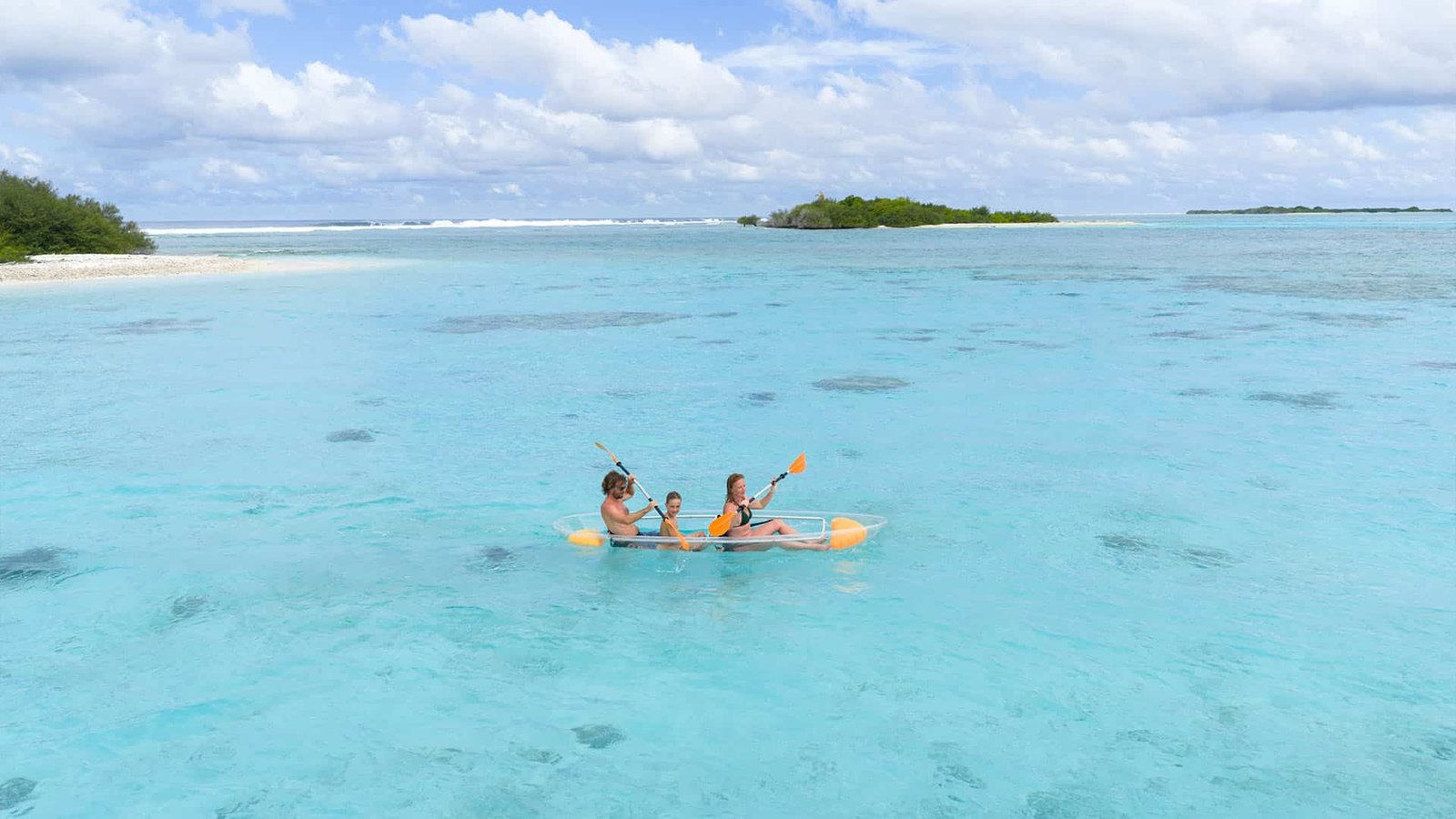 Two children standing on the wooden connecting bridge at Canareef Resort looking across the turquoise lagoon toward the neighboring local island of Hulhudhoo